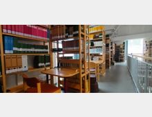 Eine Bibliothek mit hohen, hölzernen Regalen, gefüllt mit Aktenordnern, Büchern und Dokumenten. Im Vordergrund stehen Arbeitstische mit Stühlen, einer Schreibtischlampe und vereinzelt Büromaterialien; im Hintergrund verläuft ein Geländer entlang der oberen Etage.