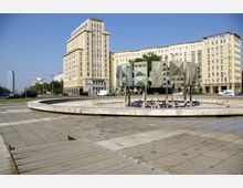 Ein Stadtplatz mit einem modernen Brunnen aus geometrischen Metallplatten im Vordergrund. Im Hintergrund befinden sich mehrstöckige Gebäude mit historischer Fassade und ein hoher, moderner Wolkenkratzer.