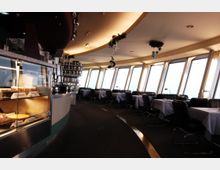 Innenansicht eines rotierenden Turmrestaurants mit großen Fensterfronten, gedeckten Tischen und moderner Einrichtung. Im Vordergrund eine beleuchtete Kuchenvitrine und eine Theke mit gestapelten Tassen und Kannen.