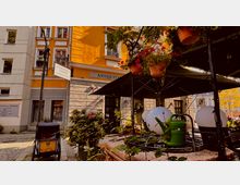 Eine städtische Szene mit einem Antiquitätengeschäft in einem gelben Gebäude. Im Vordergrund stehen Blumen, Topfpflanzen, ein Sonnenschirm und dekorative Lampen, die einen kleinen Außenbereich gestalten.