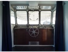 Steuerstand eines Schiffes mit einem großen Holzarmaturenbrett, zentralem Steuerrad und mehreren Kontrollinstrumenten. Dahinter befinden sich große Fenster, die den Blick nach draußen ermöglichen, eingerahmt von dunkelblauen Vorhängen.