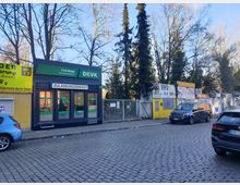 Straßenszene mit Kopfsteinpflaster, links ein kleines Bürogebäude mit grünem DEVK-Logo und der Aufschrift „Zulassungsdienst“, daneben gelbe und weiße Werbeschilder für Dienstleistungen. Im Hintergrund stehen Bäume, und am Straßenrand sind geparkte Autos zu sehen.