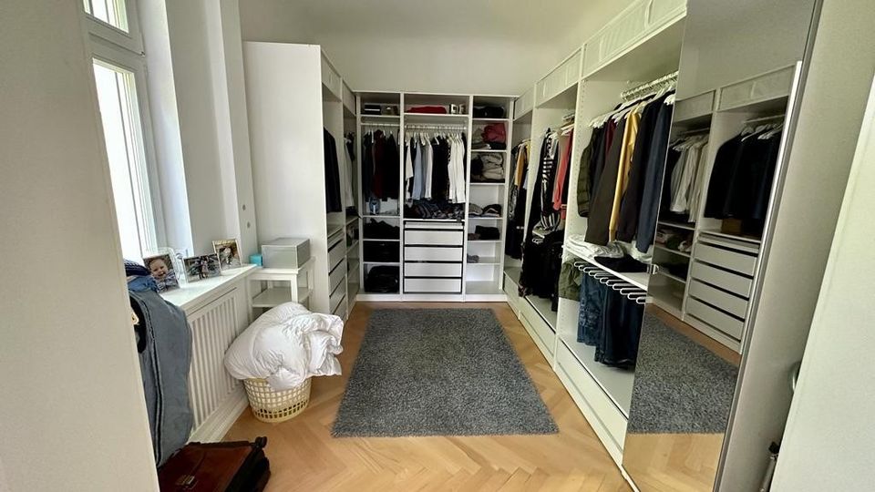 Ein begehbarer Kleiderschrank mit hölzernem Fischgrätboden, offenen Regalen, Kleiderstangen und Schubladen an beiden Seiten. Auf der rechten Seite befinden sich spiegelnde Schranktüren, und links ist ein Fenster mit einer Fensterbank, auf der persönliche Gegenstände stehen.