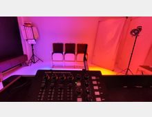 Ein Studio mit farbiger Beleuchtung (rot und violett) zeigt drei Stühle vor einer simplen weißen Kulisse. Im Vordergrund befindet sich ein Lichtsteuerpult, und im Hintergrund sind Beleuchtungselemente und Reflektoren auf Stativen erkennbar.