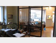 Ein Büro mit Holzboden, das durch eine Glaswand in zwei Bereiche unterteilt ist. Links steht ein Schreibtisch mit Papieren, einer Lampe und Pflanzen, rechts ein weiterer Arbeitsplatz mit einem Laptop und Schreibtischutensilien vor einem großen Fenster, das Tageslicht hereinlässt.