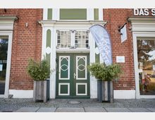 Eingang eines Gebäudes mit roter Ziegelfassade, grün-weißer zweiflügeliger Tür mit dekorativen Verzierungen und darüberliegenden Sprossenfenstern. Vor der Tür stehen zwei Holzpflanzkübel mit Bambus, und ein Schild sowie eine Fahne mit der Aufschrift "Yoga & Pilates" weisen auf ein Studio hin.