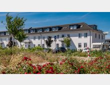 Mehrfamilienhaus mit weißer Fassade und dunklem Dach in einer Vorstadtumgebung, umgeben von Grünflächen, vereinzelten Bäumen und blühenden Sträuchern im Vordergrund. Autos und ein Wohnwagen sind in der Einfahrt des Gebäudes geparkt, bei blauem Himmel und sonnigem Wetter.