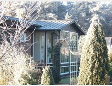 Modernes Einfamilienhaus mit einer großen verglasten Veranda, umgeben von einem Garten mit hohen Sträuchern und Bäumen. Im Hintergrund ist ein bewaldetes Gebiet sichtbar.