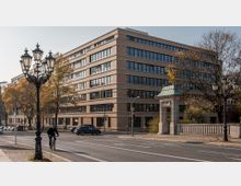Straßenszene mit einem sechsstöckigen Bürogebäude aus hellem Backstein im Hintergrund, flankiert von Bäumen mit Herbstlaub. Im Vordergrund sind eine historische Straßenlaterne, eine Radfahrerin und ein kleiner steinerner Torbogen mit grünem Dach zu sehen.
