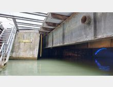 Blick auf eine überdachte Wasserpassage unter einer rostigen Beton- und Stahlkonstruktion mit sichtbaren Trägern, daneben eine Metalltreppe. Im Hintergrund leuchtet ein blauer Lichtbogen, und an der Betonwand ist ein gelbes Schild mit der Aufschrift „eisenspälterei“ befestigt.