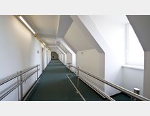 Langer, heller Flur mit schrägen Wänden und großen Fenstern auf der rechten Seite, einem grünen Teppichboden und Metallgeländern beidseits des Weges. Die Wände sind weiß, die Beleuchtung erfolgt durch eine lange Leuchtstofflampe an der Decke.