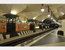 U-Bahnhof mit moderner Gestaltung, gekennzeichnet durch breite Säulen mit weißen, geometrischen Strukturen an der Decke und goldenen Lampen. Eine orangefarbene U-Bahn steht am Bahnsteig, umgeben von schwarz-weißen Kacheln und dezenten Beleuchtungselementen.