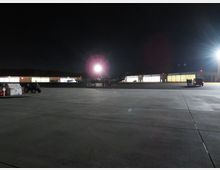 Nachtszene auf einem Flugplatz mit beleuchteten Hangars im Hintergrund. Ein Flugzeug steht auf dem Rollfeld, und eine Gruppe von Personen sowie ein Fahrzeug sind rechts im Bild erkennbar.