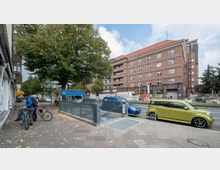 Straßenszene mit einem Eingang zur U-Bahn-Station "Afrikanische Straße", umgeben von einem großen Baum, geparkten Autos und einem mehrstöckigen Backsteingebäude mit Baustellenbereich im Hintergrund. Auf dem Gehweg stehen zwei Personen mit Fahrrädern.