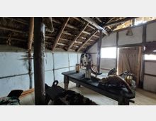 Ein rustikaler Innenraum mit einem Tisch aus Holz, auf dem Dekorationen wie ein Trinkhorn, Laternen und Tierfelle liegen. Die Wände sind mit Lehm ausgefacht, das Dach besteht aus sichtbaren Holzbalken und Stroh, und ein hölzernes Tor sowie ein Fenster lassen Tageslicht herein.