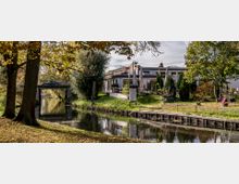 Uferlandschaft mit einem kleinen Fluss, gesäumt von Bäumen und Grünflächen. Im Hintergrund ein modernes Gebäude mit Terrasse und Sonnenschirmen sowie rechts eine Brücke, über die ein Auto fährt.