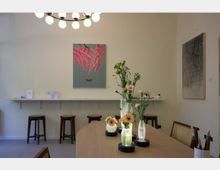 Minimalistisch eingerichteter Innenraum mit einem großen Holztisch im Vordergrund, auf dem dekorative Blumenvasen stehen. Im Hintergrund eine Wand mit zwei Kunstwerken, einer schmalen weißen Ablagefläche und mehreren Holzhockern.