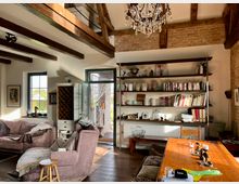 Wohnraum in einem rustikalen Stil mit sichtbaren Holzbalken und Backsteinwänden. Der Raum enthält eine Sitzecke mit einem Sofa, mehrere Bücherregale, einen Holztisch mit Stühlen und eine Glastür, die nach außen auf eine Terrasse führt.