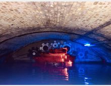 Ein unterirdischer Tunnel mit gewölbtem Mauerwerk, dessen Wände mit Graffiti von Zahnrädern bemalt sind; Wasser bedeckt den Boden und bunte Lichter in Blau und Rot beleuchten den Raum.