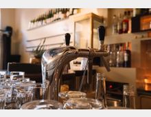 Nahaufnahme eines Tresens in einer Bar mit einem glänzenden Bierzapfhahn im Vordergrund. Im Hintergrund sind leere Flaschen, Gläser und eine Regalwand mit aufgereihten Getränkeflaschen zu sehen.