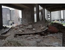 Industriegelände mit verfallenem Gebäude, Betonpfeilern und Schutthaufen im Vordergrund. Im Hintergrund sind große, runde Betonsilos und einsturzgefährdete Strukturen sichtbar.