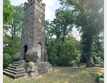 Steinerner Turm aus unregelmäßigem Mauerwerk mit quadratischem Grundriss und Zinnen, umgeben von großen Felsblöcken und einer kleinen Treppe, inmitten eines waldreichen Parks mit Rasenfläche und hohen Bäumen.