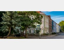 Straßenszene mit historischen Wohnhäusern in verschiedenen Pastellfarben, darunter grün, weiß und blau, entlang einer Kopfsteinpflasterstraße; im Vordergrund stehen mehrere Bäume, darunter ein Nadelbaum, sowie ein Straßenschild mit der Aufschrift „Elmshorner Platz“.