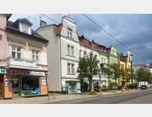 Straßenansicht mit mehrstöckigen Wohnhäusern, die in verschiedenen Farben gestrichen sind, darunter Weiß, Grün und Gelb, mit dekorativen Fassadenelementen. Im Vordergrund befinden sich ein Gehweg, Bäume sowie eine Bushaltestelle mit Sitzbank, daneben ein kleines Geschäft mit der Aufschrift „Garten Center“.