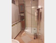 Badezimmer mit einer halbrunden Duschkabine aus Glas, einer beige gefliesten Wand und einem Handtuchhalter an der rechten Seite. Links befindet sich ein Heizkörper unter einem Fenster sowie ein Handtuchschrank aus Holz.