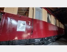 Roter, glänzender historischer Eisenbahnwaggon mit beigem oberen Bereich, Fenstern mit Vorhängen und einer Informationstafel an der Tür. Der Waggon steht in einem überdachten Ausstellungsraum mit Holzdach.