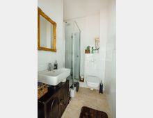 Kleines Badezimmer mit weißen Fliesen an den Wänden und beigefarbenen Fliesen auf dem Boden. Links befinden sich ein rechteckiges Waschbecken auf einem dunklen Holzschrank und ein goldgerahmter Spiegel darüber, rechts eine Glasduschkabine, eine Wandtoilette und dekorative Accessoires wie eine Orchidee.