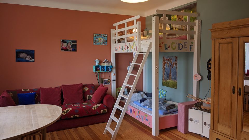 Kinderzimmer mit Etagenbett aus hellem Holz und Leiter, daneben ein rotes Sofa und ein runder Tisch auf Holzdielenboden. Farbige Wände mit Bildern und ein großer Holzschrank prägen den Raum.