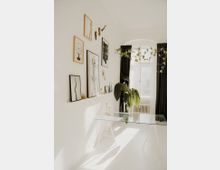 Helles, minimalistisch eingerichtetes Zimmer mit einer weißen Wand, an der gerahmte Bilder und Dekorationen hängen. Im Vordergrund steht ein Glastisch mit zarten, goldenen Kerzenhaltern, während eine große grüne Pflanze neben einem Fenster mit schwarzen Vorhängen steht.