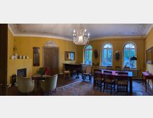 Ein historisch eingerichteter Raum mit gelben Wänden, einer Holztür und hohen, bogenförmigen Fenstern. Die Ausstattung umfasst einen Kronleuchter, einen Flügel, eine Sitzgruppe mit antiken Sesseln und einen Esstisch mit Stühlen, ergänzt durch dekorative Elemente wie Spiegel und Keramiken.
