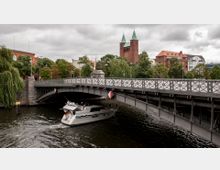 Eine Brücke über einen Fluss mit einem Metallgeländer und Verzierungen, darunter ein vorbeifahrendes Motorboot. Im Hintergrund sind Bäume, ein historisches Backsteingebäude und eine Kirche mit zwei Türmen und grünen Dächern zu sehen.