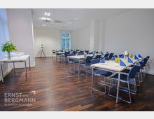 Ein moderner, heller Veranstaltungsraum mit Holzboden, weißen Wänden und deckenhohen Fenstern. Rechteckige Tische mit blauen Stühlen sind ordentlich gedeckt, mit blauen Stoffservietten, Gläsern und Blumendekoration.