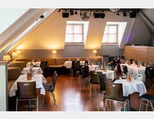 Ein Restaurant- oder Veranstaltungsraum mit Dachschrägen, gedeckten Tischen mit weißen Tischdecken, Stühlen aus Holz und Ledersitzgruppen an den Wänden. Warmes Lampenlicht und zwei Fenster mit Holzjalousien sorgen für eine gemütliche Atmosphäre.