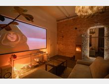 Gemütliches Wohnzimmer mit Backsteinwand und warmem Licht, ausgestattet mit einem Beamer, einem Kamin und modernen Möbeln. Ein großer Spiegel reflektiert den Raum, und eine Hängelampe sorgt für zusätzliche Beleuchtung.