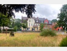 Städtische Szene mit mehrstöckigen Wohn- und Geschäftshäusern mit Sattel- und Mansarddächern im Hintergrund. Im Vordergrund befindet sich eine Wiese mit hohem Gras sowie eine kleine steinerne Skulptur, umgeben von einzelnen Bäumen.