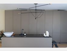 Moderne Küche mit einer zentralen Kücheninsel in Grautönen, schwarzer Arbeitsfläche, integriertem Waschbecken und Dekorationsobjekten wie einer Keramikschale und Kerzen. Im Hintergrund befinden sich glatte, wandhohe Schränke in einer minimalistischen Gestaltung.