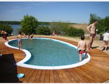 Kinder spielen in einem kleinen, nierenförmigen Pool, der von einer Terrasse aus Holz umgeben ist. Im Hintergrund sind ein See, Büsche und Bäume unter einem klaren blauen Himmel zu sehen.
