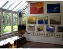 Künstleratelier mit großem Fenster und Glasdach, das einen Blick auf einen grünen Garten bietet. An der Innenwand hängen zahlreiche gerahmte Bilder in unterschiedlichen Größen und Farben, darunter Landschaftsdarstellungen und abstrakte Kunstwerke. Ein Arbeitstisch mit Malutensilien steht im Vordergrund.