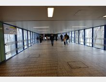 Ein langer, überdachter Fußgängerkorridor mit großen Fenstern an beiden Seiten, die mit blauen Rahmen eingefasst sind. Menschen gehen in beide Richtungen, während ein Schild die U-Bahn-Linie U7 anzeigt.