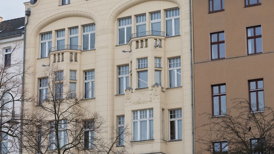 Mehrstöckiges helles Wohn- und Geschäftshaus mit geschwungenem Giebel und rotem Ziegeldach, vielen hohen Fenstern und kleinen Balkonen; im Erdgeschoss ein Durchgang mit der Aufschrift „Engelbecken-Hof“. Davor stehen kahle Bäume und eine niedrige Mauer mit Graffiti an einer Straßenkante.