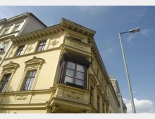 Gelbes historisches Gebäude mit aufwendigen Fassadendetails wie Zierornamenten und verzierten Fenstern. Ein dekorativer Erker mit schwarzem Geländer ragt hervor, daneben ein moderner Straßenlaternenmast vor blauem Himmel.