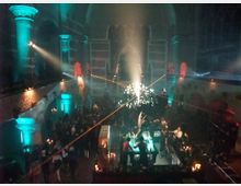 Innenraum einer beleuchteten Kirche, die als Veranstaltungsort genutzt wird. Es gibt eine Bar im Vordergrund, zahlreiche Gäste, bunte Lichtstrahler, sowie Dekoration und Architektur im gotischen Stil.