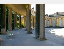 Halbrunde Kolonnade vor einem historischen gelben Gebäude mit hohen Fenstern. Die steinernen Säulen sind reich verziert und umgeben einen gepflasterten Hof; im Hintergrund sind Grünflächen zu sehen.