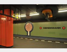 U-Bahnhof Siemensdamm in Berlin mit grüner Wandgestaltung und geometrischen Elementen in Blau und Rot; ein großer roter Pfeiler steht links im Bild, darüber verlaufen Deckenleuchten und dunkle Paneele.