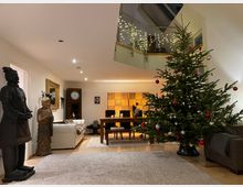Wohnzimmer mit festlich geschmücktem Weihnachtsbaum rechts und asiatisch inspirierten Statuen links. Im Hintergrund befinden sich ein Esstisch, ein Sofa, eine Wanduhr sowie eine Galerie mit Glasgeländer, illuminiert durch Lichterketten.