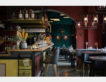 Modernes Restaurant mit einer stilvollen Einrichtung, darunter eine lange Bar mit Hockern, hängenden Gläsern und Dekorationen wie gelben Blumen in einer Vase. Im Hintergrund befinden sich gedeckte Tische mit Blumenarrangements, dunkle Wände in Grün- und Rottönen sowie moderne Pendelleuchten.